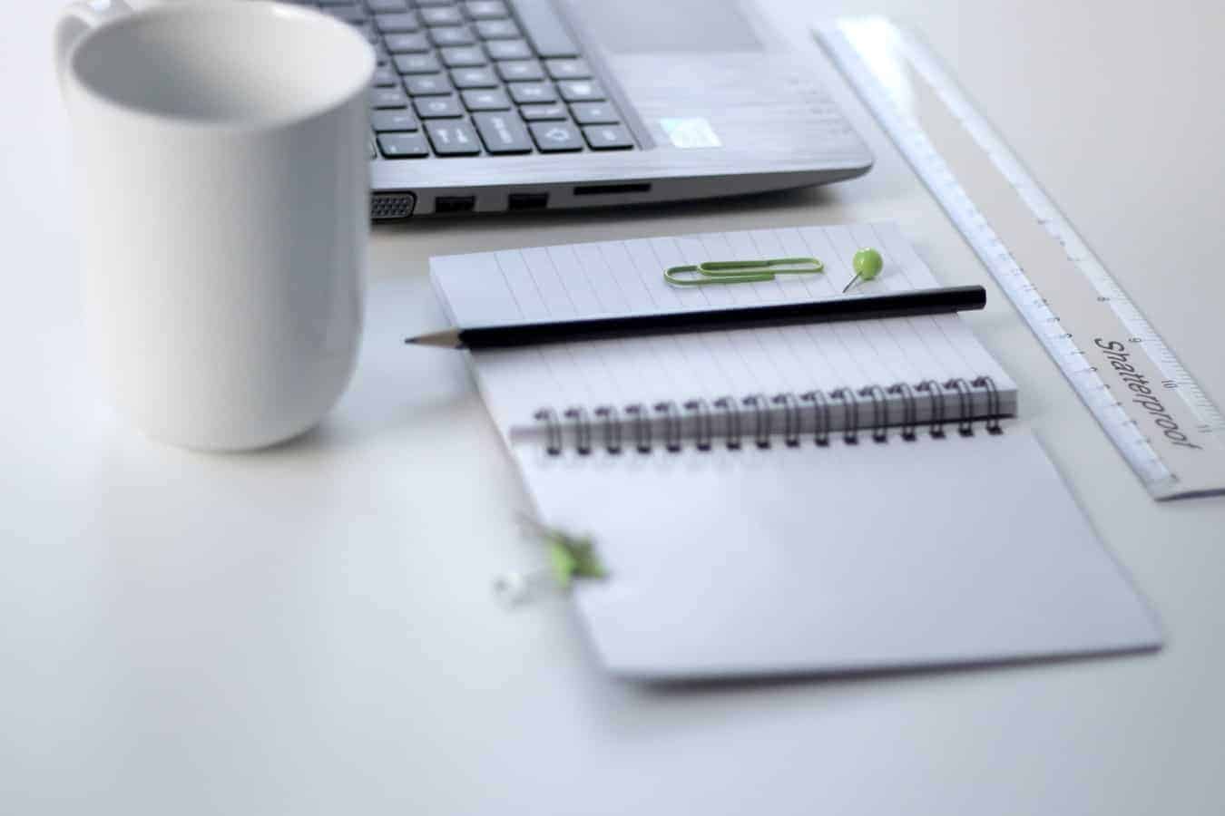 A white desk with an open notebook, white mug, white laptop and clear ruler