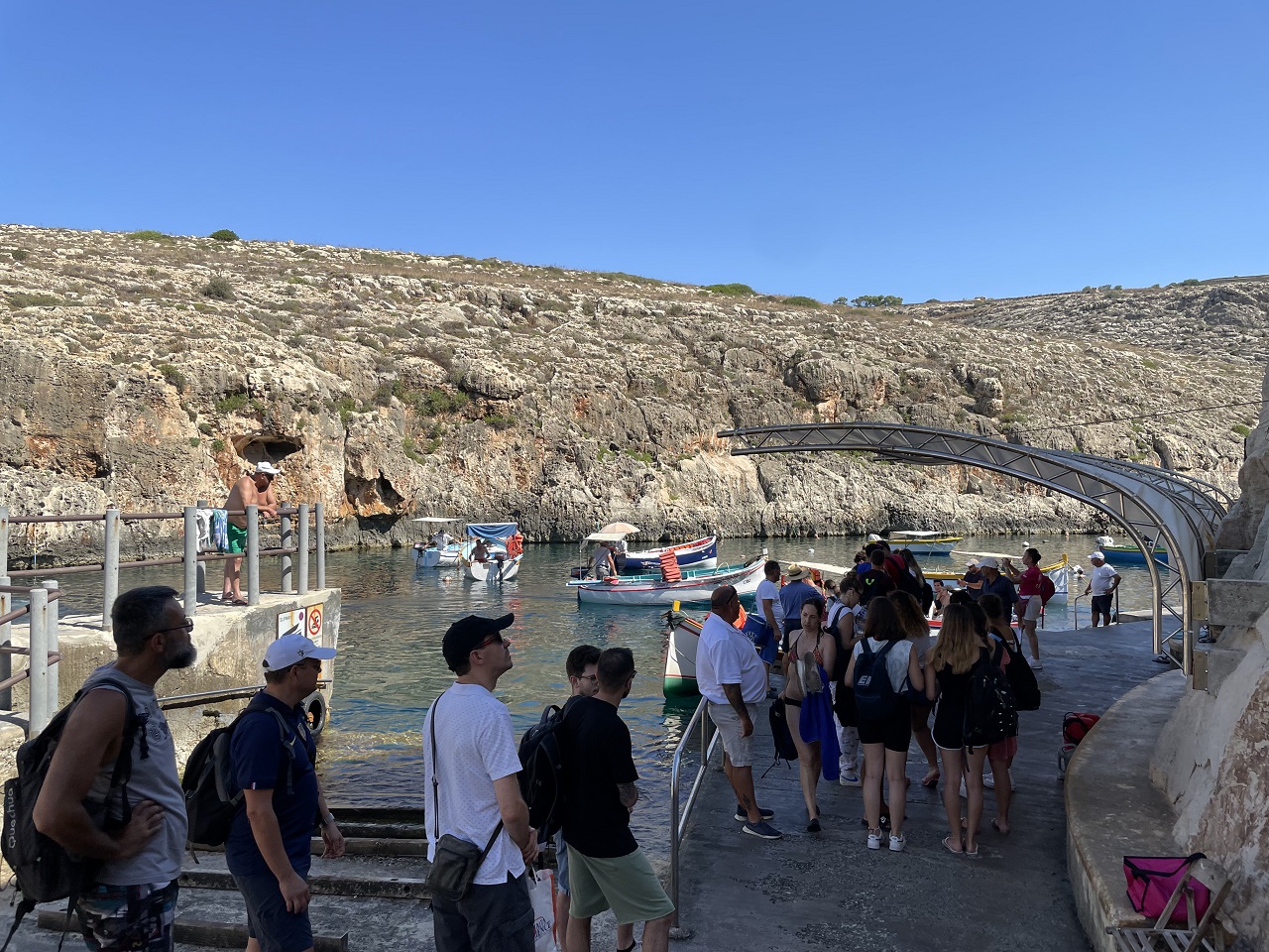 fifteen people in line waiting to board the blue grotto boats in malta with boats and cliffs in background