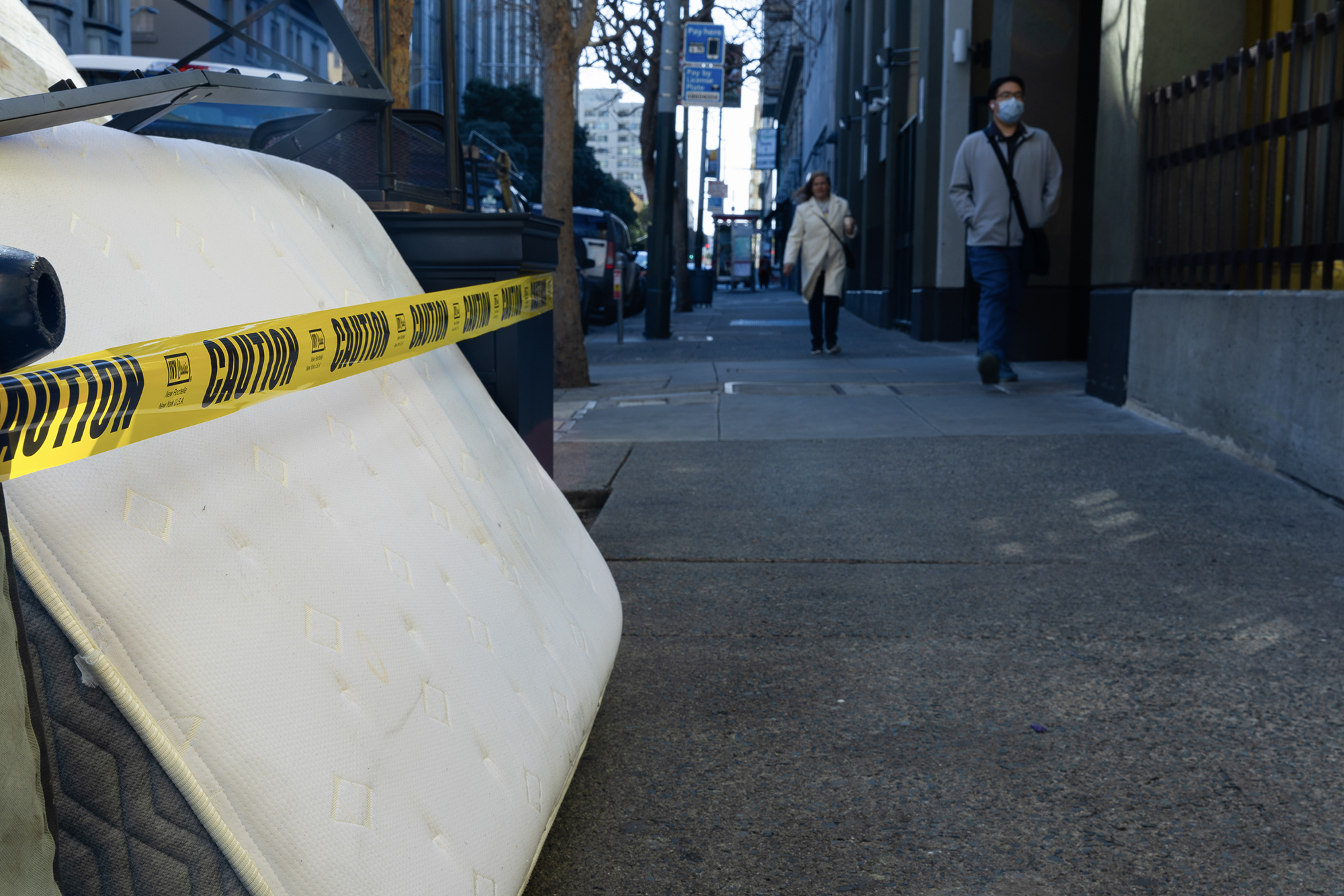 A discarded mattress leans against a pile of furniture on a city sidewalk, partially wrapped in yellow caution tape. In the background, two people walk past a building with dark green and yellow walls, one wearing a face mask and carrying a shoulder bag. The urban street is lined with parked cars, trees, and tall buildings, with shadows cast by the afternoon light.