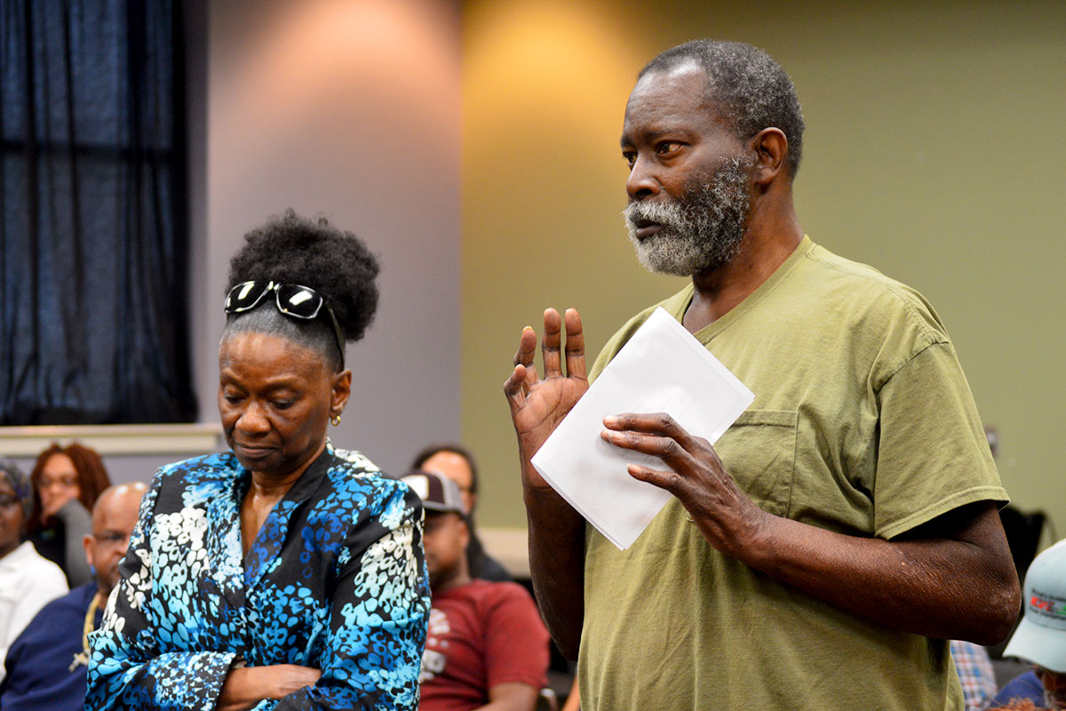 A woman and a man stand among a seated audience, talking towards the front of the room